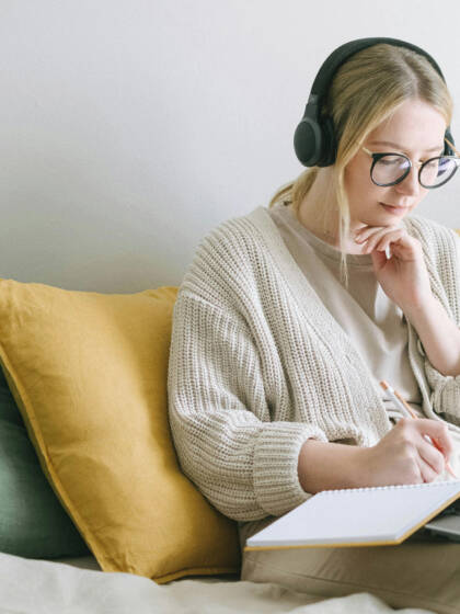A girl is sitting on a sofa and workin on her laptop. She has headphones on and she is writing on paper.