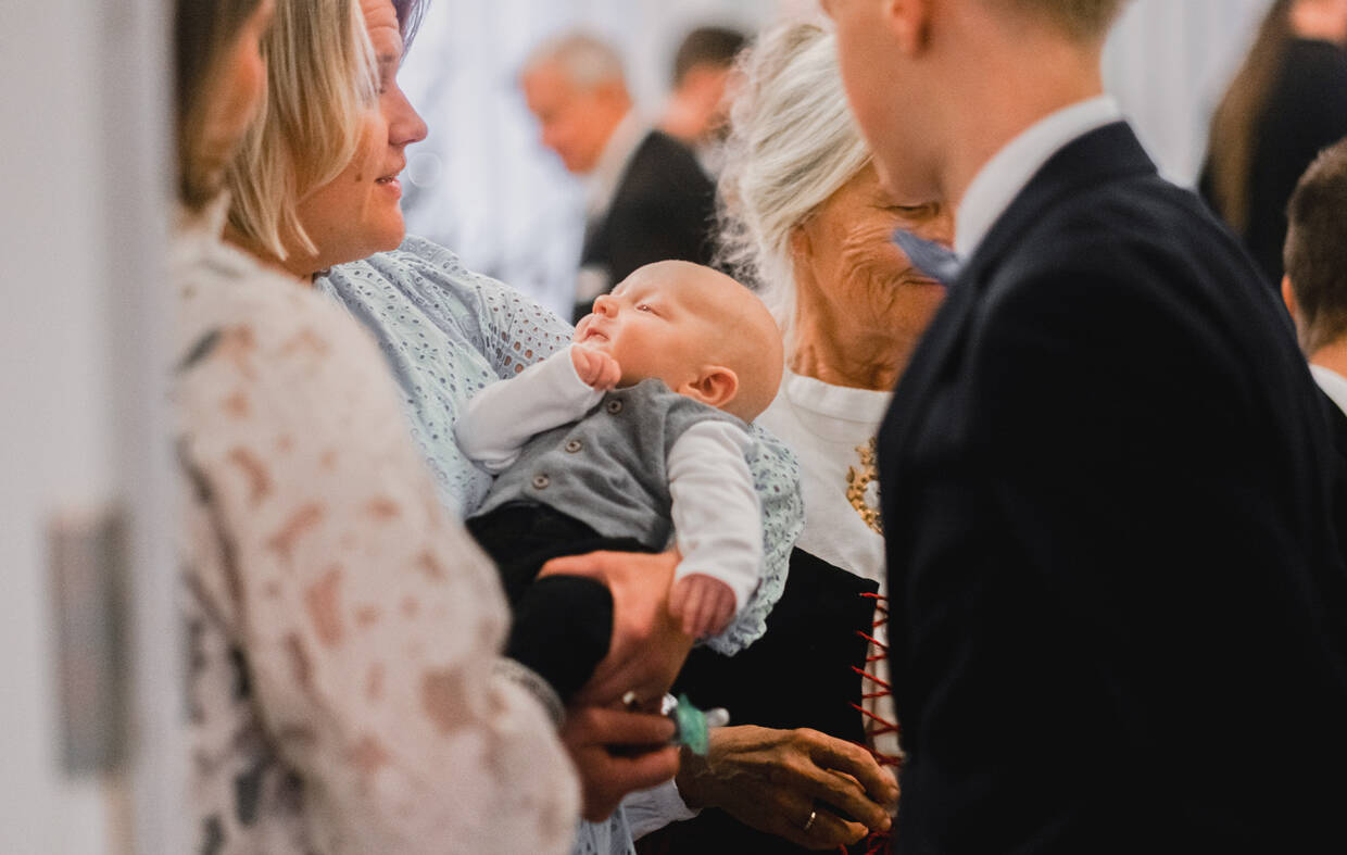 A baby is in the woman's arms, where the christening guests can admire him.