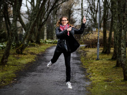 Polku ja ihminen joka hyppää ilmaan. Hänellä on tummat vaatteet ja pinkkiä kaulahuivissa. Ympärillä nurmikkoa ja puita, joissa ei ole lehtiä.