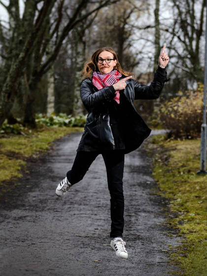 Polku ja ihminen joka hyppää ilmaan. Hänellä on tummat vaatteet ja pinkkiä kaulahuivissa. Ympärillä nurmikkoa ja puita, joissa ei ole lehtiä.