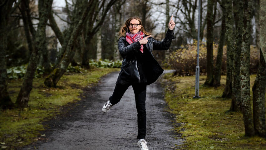 Polku ja ihminen joka hyppää ilmaan. Hänellä on tummat vaatteet ja pinkkiä kaulahuivissa. Ympärillä nurmikkoa ja puita, joissa ei ole lehtiä.