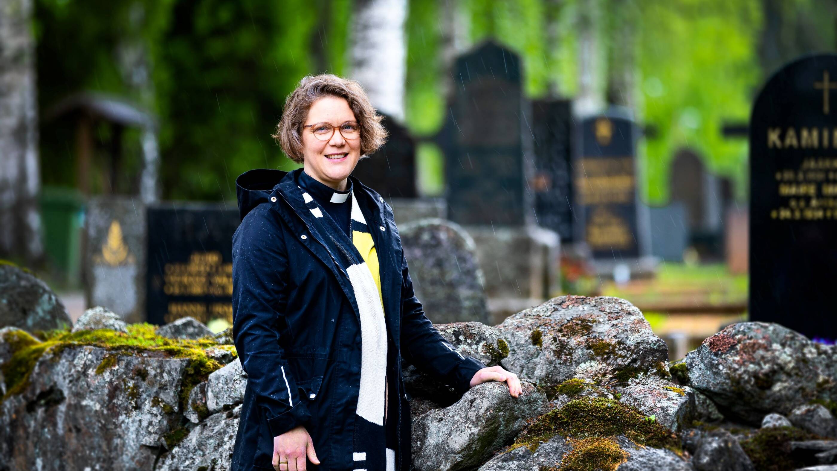 Naispapi nojaa kivimuuriin hautausmaalla. Taustalla hautakiviä.