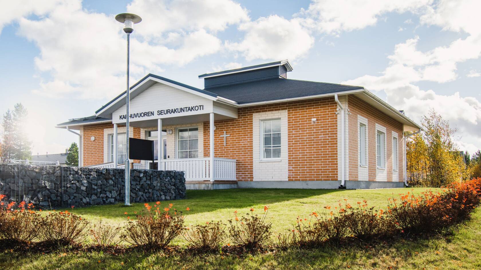Keltatiilinen rakennus, jossa harmaa katto ja valkoiset ovet ja ikkunan karmit. Ulko-oven edessä katos. Oven edessä kivimuuri ja ympärillä nurmikkoa ja lehtipensaita.