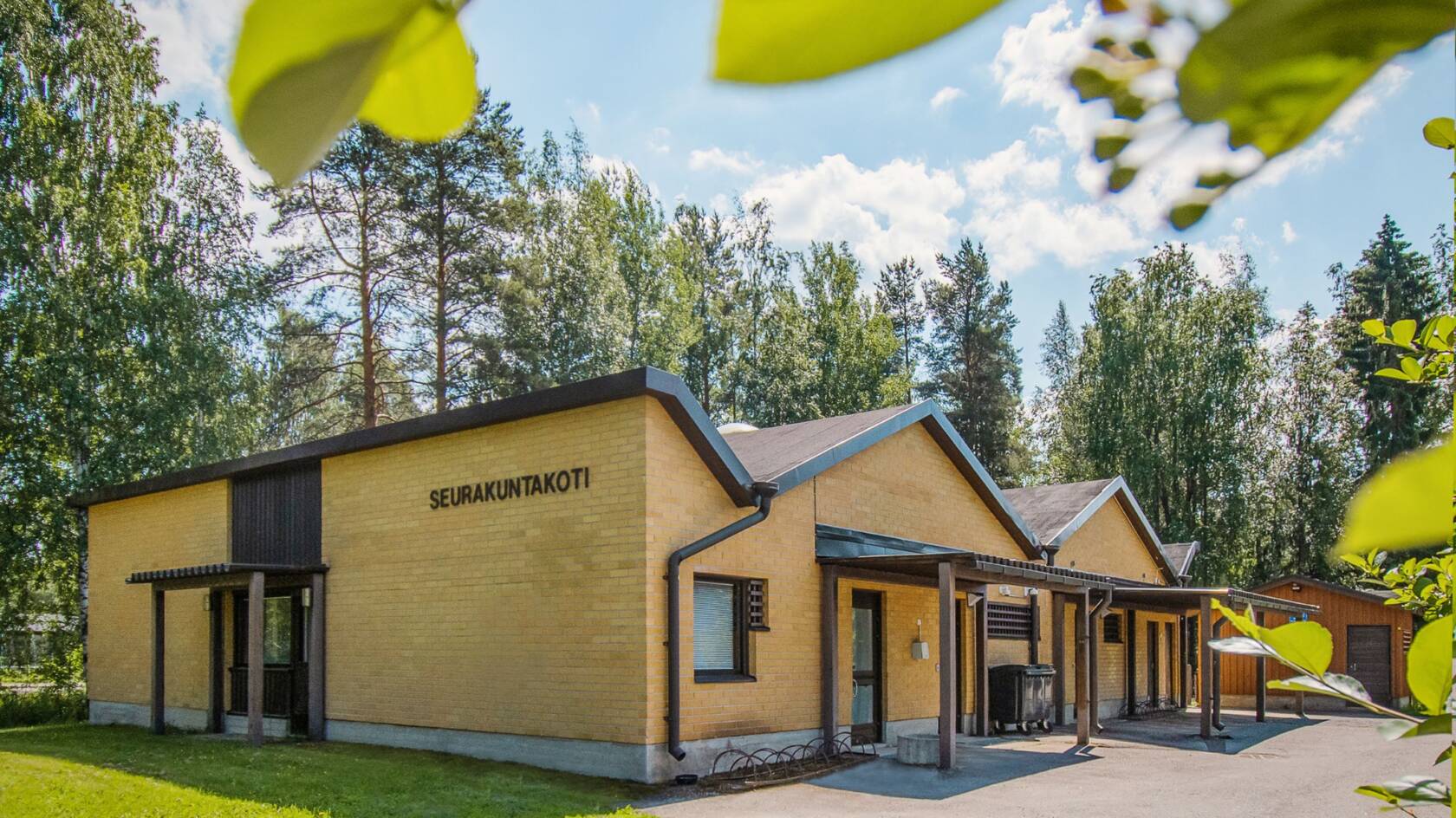 Keltatiilinen takennus ruskeilla ikkunakarmeilla ja ovilla. Ympärillä nurmikkoa. Kuvan etualalla puun lehtiä.