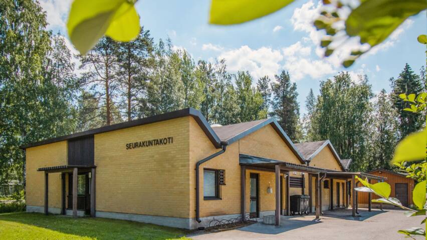 Keltatiilinen takennus ruskeilla ikkunakarmeilla ja ovilla. Ympärillä nurmikkoa. Kuvan etualalla puun lehtiä.
