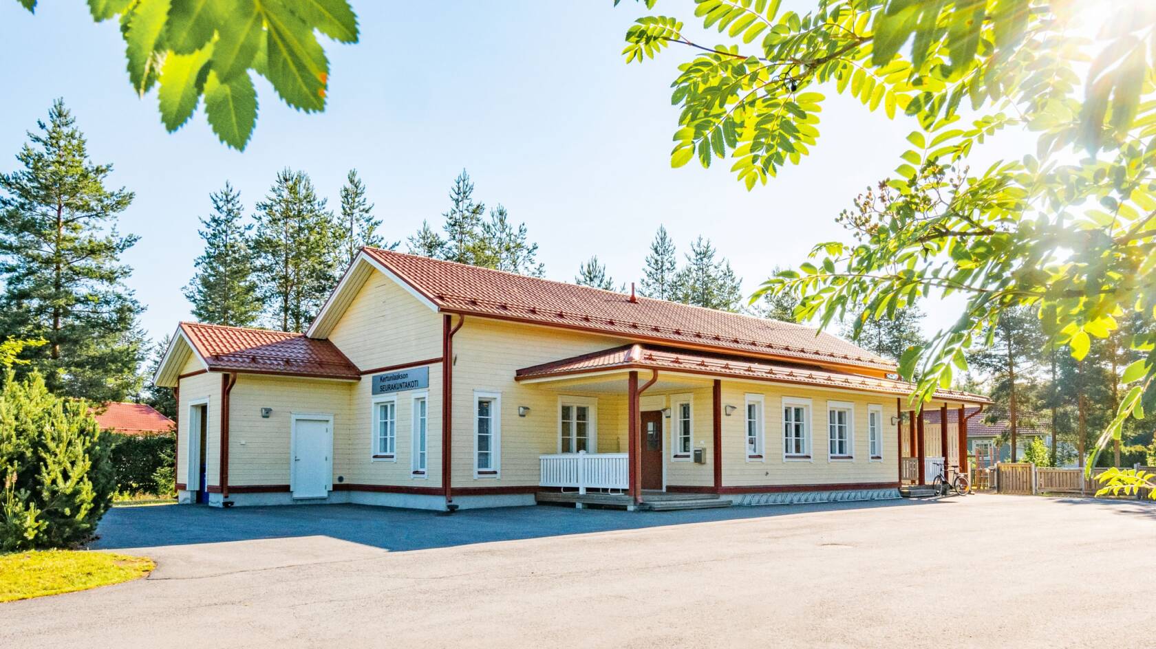 Vaalean keltainen rakennus, jossa punapeltinen katto. Valkoiset ovet ja karmit ikkunoissa. Suuri hiekkapiha ja ympärillä puita. Kuvan etualalla näkyy pihlajan oksia.