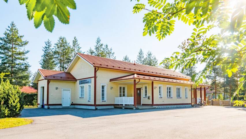 Vaalean keltainen rakennus, jossa punapeltinen katto. Valkoiset ovet ja karmit ikkunoissa. Suuri hiekkapiha ja ympärillä puita. Kuvan etualalla näkyy pihlajan oksia.