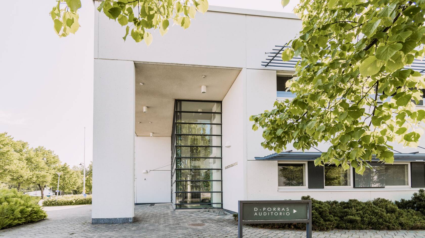 Valkoinen kivirakennus, jossa paljon ikkunapintaa. Kyltti D-ovi auditorio. Rakennuksen edessä laatoitusta, asfalttia ja nurmikkoa. Kuvassa etualalla lehtipuun oksa.