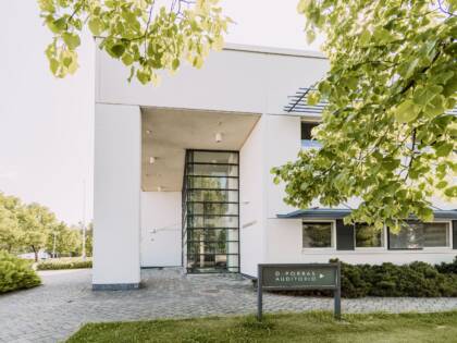 A white stone building with plenty of windows. A sign reading ‘Door D – Auditorium’. In front of the building there is paving, tarmac and a lawn. In the foreground of the picture is a branch of a deciduous tree.