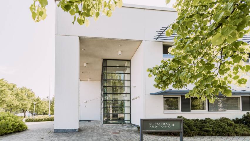 Valkoinen kivirakennus, jossa paljon ikkunapintaa. Kyltti D-ovi auditorio. Rakennuksen edessä laatoitusta, asfalttia ja nurmikkoa. Kuvassa etualalla lehtipuun oksa.
