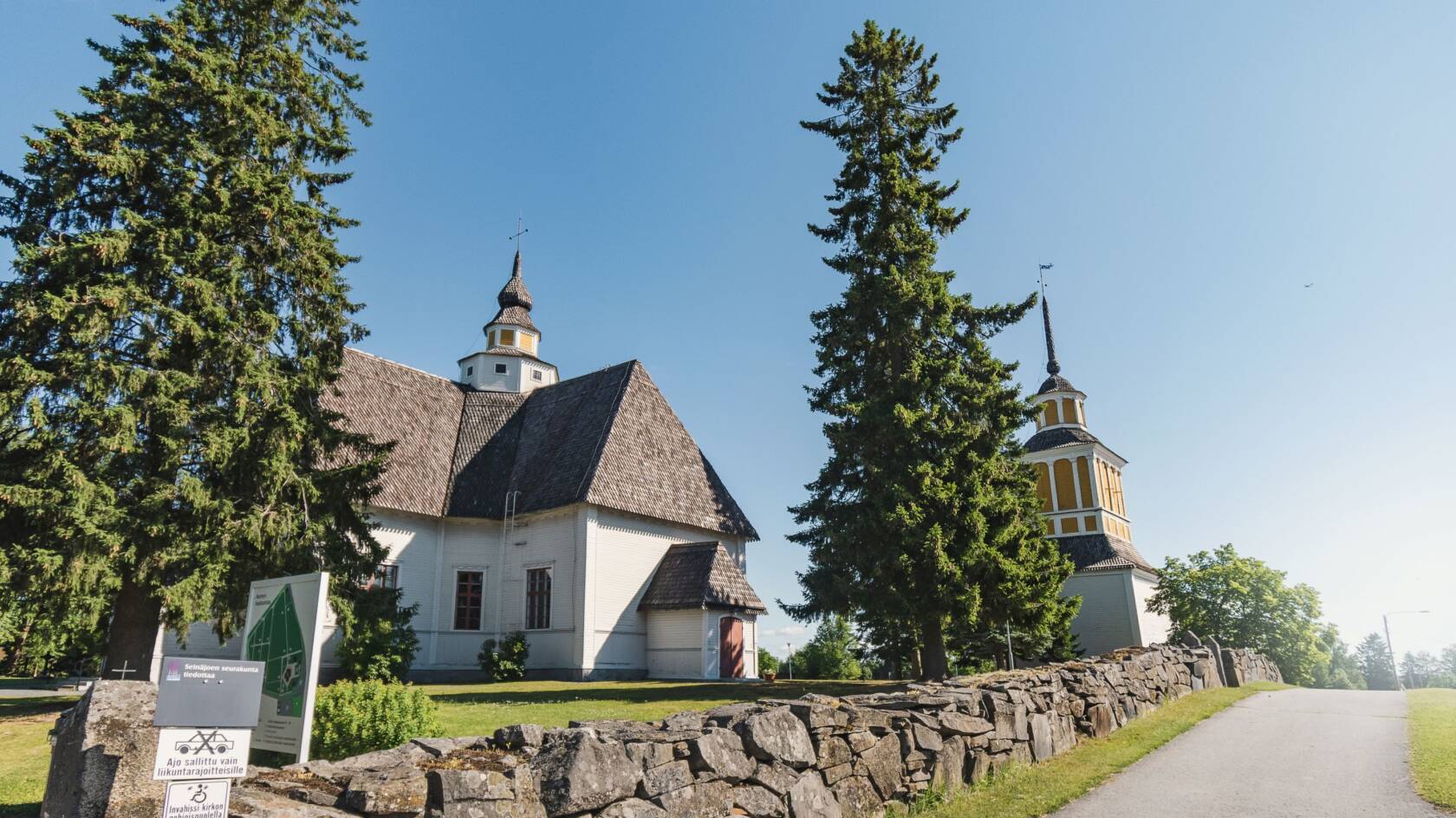 Valkoinen kirkko ruskealla katolla, keltaisella tornilla. Kirkon vieressä kaksi isoa puuta ja kivimuuri.