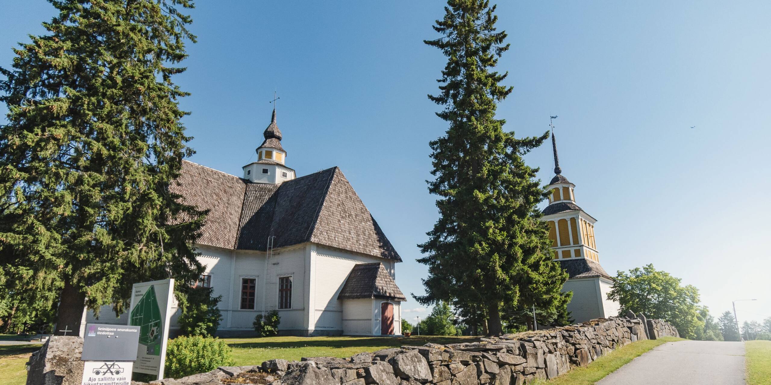 Valkoinen kirkko ruskealla katolla, keltaisella tornilla. Kirkon vieressä kaksi isoa puuta ja kivimuuri.