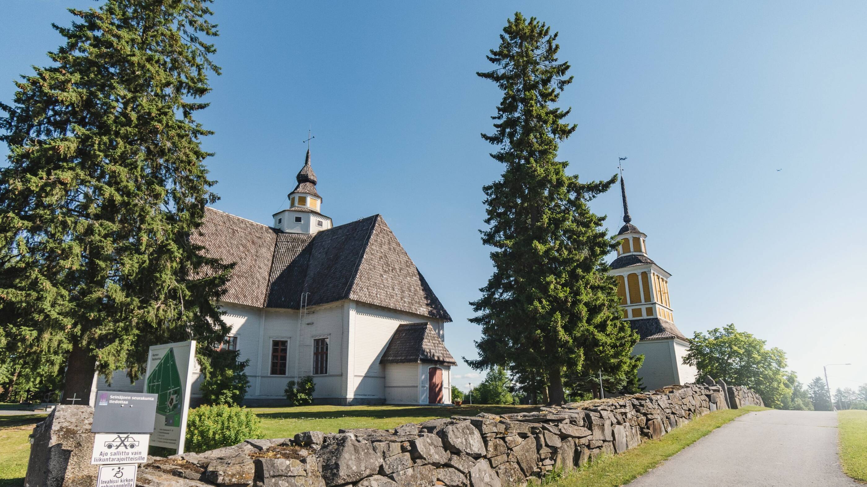 Valkoinen kirkko ruskealla katolla, keltaisella tornilla. Kirkon vieressä kaksi isoa puuta ja kivimuuri.