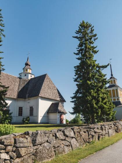 Valkoinen kirkko ruskealla katolla, keltaisella tornilla. Kirkon vieressä kaksi isoa puuta ja kivimuuri.