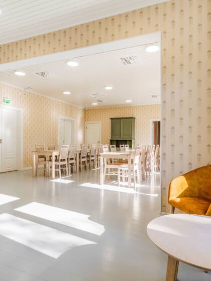 A room with a view into another room. Light-colored floor, yellow patterned wallpaper. In the foreground of the photo, an orange armchair and a round, low table. In the background of the photo, a dining room with dining sets and a green sideboard.