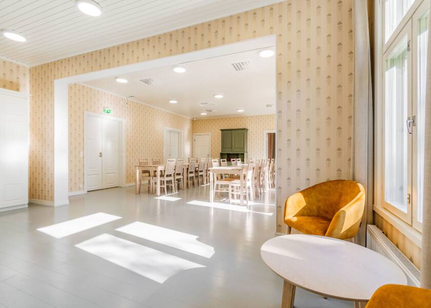 Tila, josta näkyy toiseen huoneeseen. Vaalea lattia, keltainen kuviollinen tapetti. Kuvan etualalla oranssi nojatuoli ja pyöreä matala pöytä. Kuvassa takana sali, jossa ruokailuryhmiä. ja vihreä senkki.