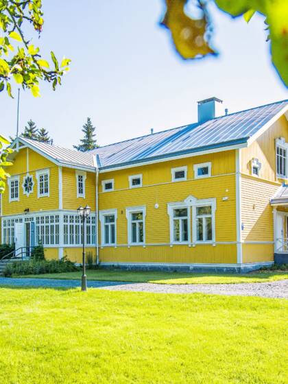Keltainen vanha hirsirakennus, jossa valkoiset ikkunanpielet ja lasikuisti. paljon nurmikkoa, hiekkapiha ja puita ympärillä.