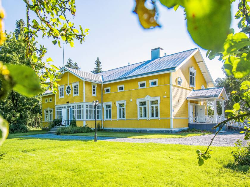 Keltainen vanha hirsirakennus, jossa valkoiset ikkunanpielet ja lasikuisti. paljon nurmikkoa, hiekkapiha ja puita ympärillä.