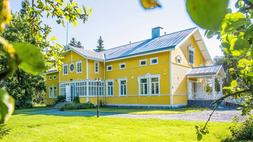 Keltainen vanha hirsirakennus, jossa valkoiset ikkunanpielet ja lasikuisti. paljon nurmikkoa, hiekkapiha ja puita ympärillä.