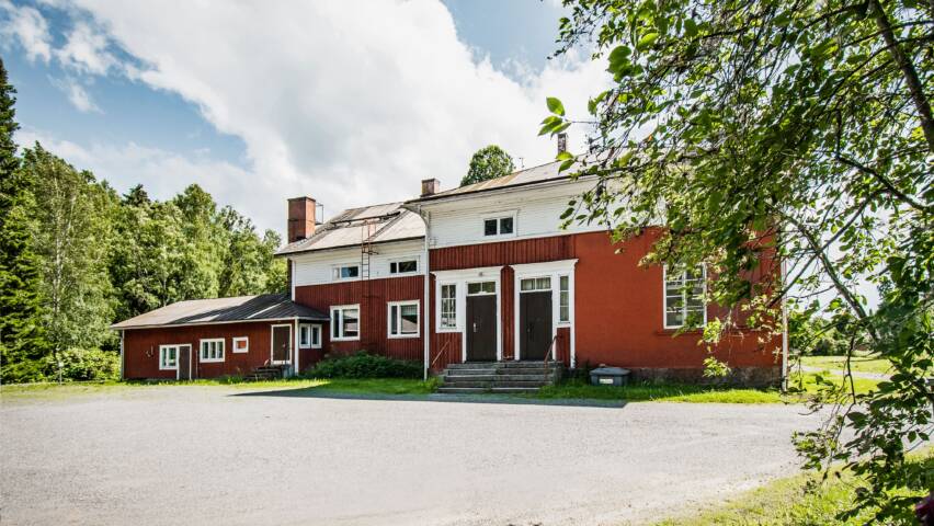 Punainen kaksikerroksinen puutalo, piha, jonka ympärillä metsikköä.