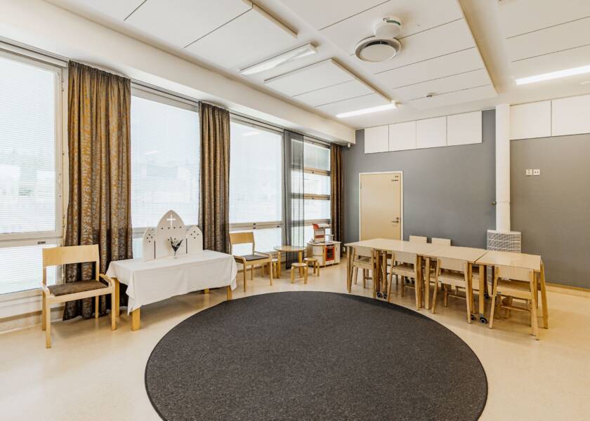 Tila, jossa vaalea lattia ja isot ikkunat. Keskellä harmaa matto ja pöytäryhmä. Ikkunan edessä pieni alttari. Ruskeanharmaapohjaiset kuviolliset verhot ja harmaa takaseinä.