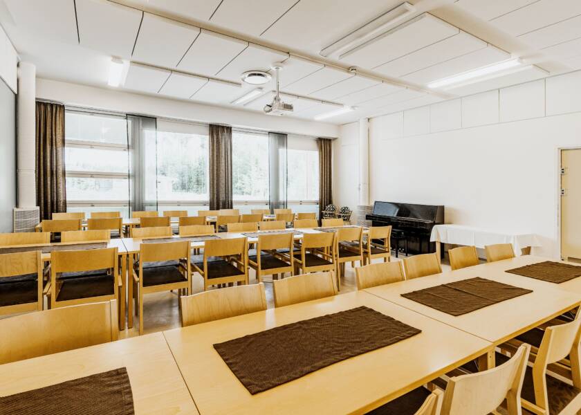 Tila, jossa isot ikkunat ja vaalean puun värisiä pöytäryhmiä. Ikkunan edessä pieni alttari. Ruskeanharmaapohjaiset kuviolliset verhot ja musta piano. Ruskeat tabletit pöydissä.