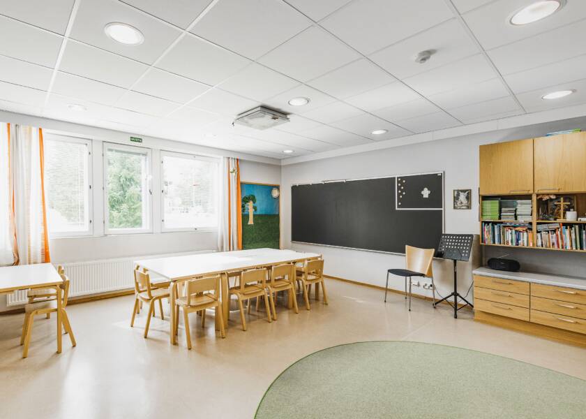 Seinällä harmaa liitutaulu ja sen vieressä kirjahylly. Vihreä pyöreä matto ja vaalean puun värisiä lasten kalusteita. Ikkuna ja vaaleat verhot, joissa hieman oranssia raitaa.