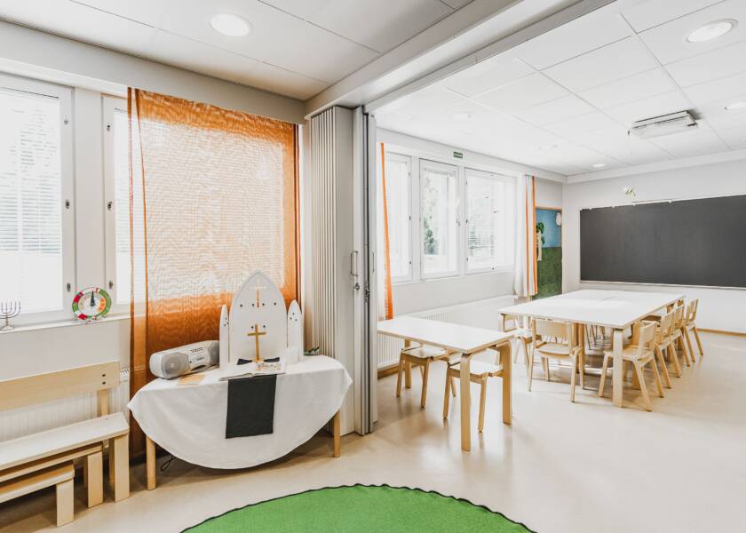 Kuvassa kaksi tilaa, jotka saadaan erotettua haitariovella. Takana näkyy harmaa liitytaulu ja lasten kalusteita pöytiä ja tuoleja. Kuvassa etualalla ikkunoita ja oranssi verho, sekä ruohonvihreä matto ja pieni alttari sekä musiikkisoitin.