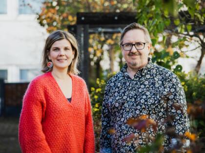 Nainen ja mies seisovat vierekkäin. Taustalla syksyinen maisema. naisella oranssi paita ja miehellä sininen kukallinen paita.