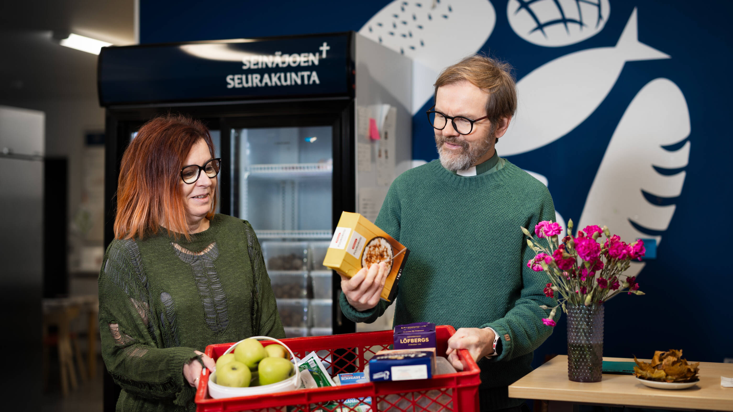 Ruokapankin logo sinisellä seinällä, jossa leipää ja kaloja. Kuvassa nainen ja mies, joilla ruokakori käsissään.