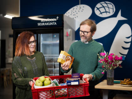 Ruokapankin logo sinisellä seinällä, jossa leipää ja kaloja. Kuvassa nainen ja mies, joilla ruokakori käsissään.