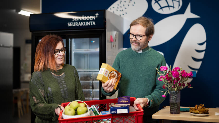 Ruokapankin logo sinisellä seinällä, jossa leipää ja kaloja. Kuvassa nainen ja mies, joilla ruokakori käsissään.