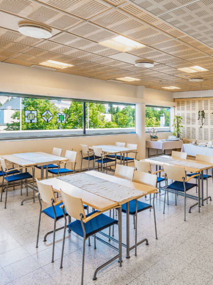 A room with light-colored wood, rectangular tables, each with four chairs upholstered in blue fabric.