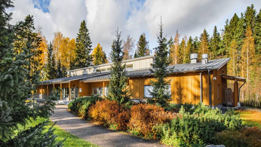 Keltainen rakennus, jonka edessä kulkee jalankulkutie, joka näyttää olulta. Ympärillä kuusia ja pensaita. KUva kuvattu syksyllä ruskan aikaan.
