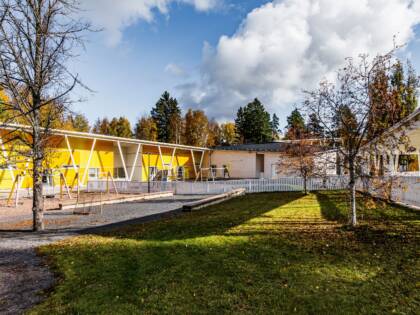 Yellow building with a playground in front. A lawn and a white fence surround the area. One section of the building is made of light-coloured brick.