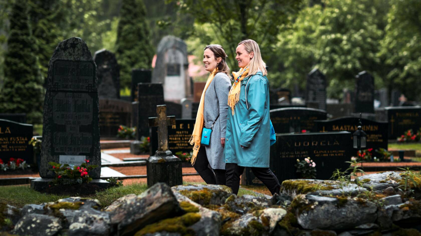 Kaksi naista kävelee hautausmaalla. Toisella naisella sininen takki ja vaaleta hiukset ja toisella naisella hat,aa takki ja ruskeat hiukset. Molemmilla keltaiset huivit. Takana näkyy vihreitä pensaita ja puita, sekä hautakiviä ja kuvan etualalla vanha kivimuuri.