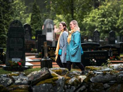 Two women are walking in a cemetery. One woman is wearing a blue coat and has fair hair, whilst the other is wearing a hat, a coat and has brown hair. Both are wearing yellow scarves. In the background, there are green bushes and trees, as well as gravestones, and in the foreground of the picture there is an old stone wall.