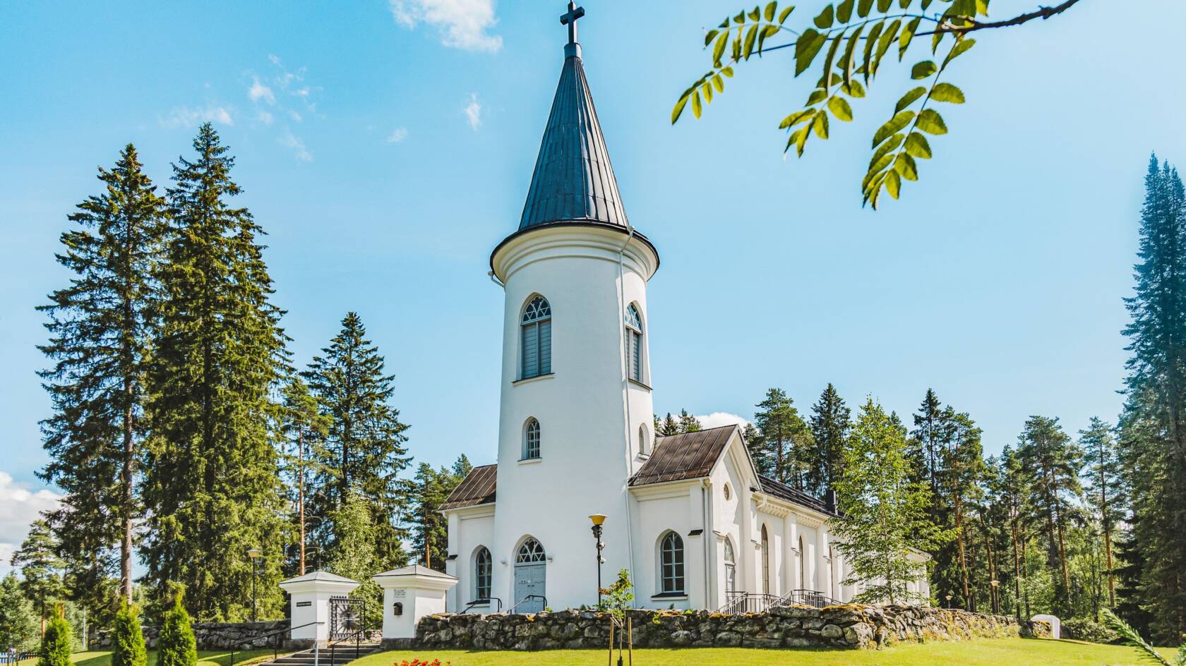 Valkoinen kirkko, vihreää nurmikkoa ja puita ympärillä. Etualalla pihlajan oksa.