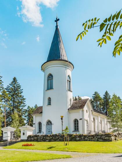 Valkoinen kirkko, vihreää nurmikkoa ja puita ympärillä. Etualalla pihlajan oksa.