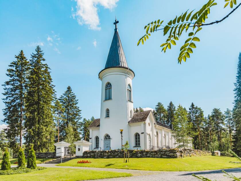 Valkoinen kirkko, vihreää nurmikkoa ja puita ympärillä. Etualalla pihlajan oksa.