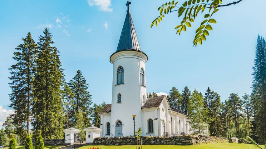 Valkoinen kirkko, vihreää nurmikkoa ja puita ympärillä. Etualalla pihlajan oksa.