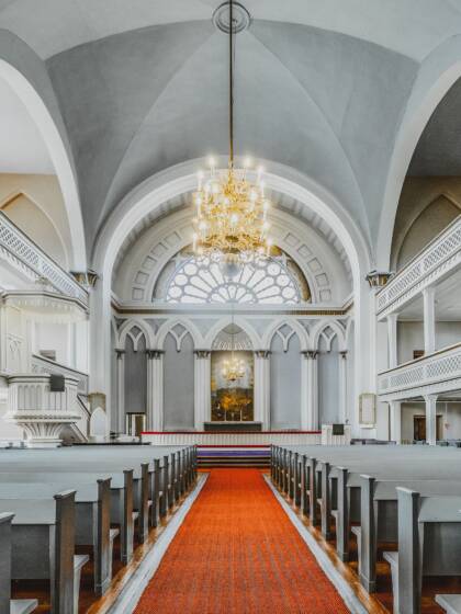 A church aisle with grey pews on both sides, an altar at the front, and a chandelier hanging from the ceiling. A red carpet runs down the aisle. The church also features galleries and vaulted arches.