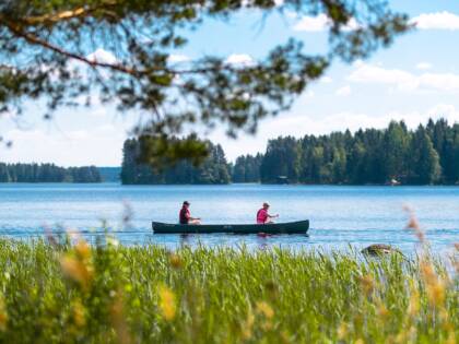 Vene, jossa kaksi ihmistä järvellä. Etualalla kaislikkoa, puita ja järven takana puita.