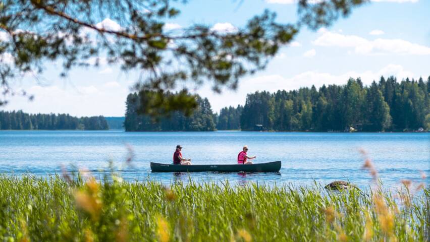 Vene, jossa kaksi ihmistä järvellä. Etualalla kaislikkoa, puita ja järven takana puita.