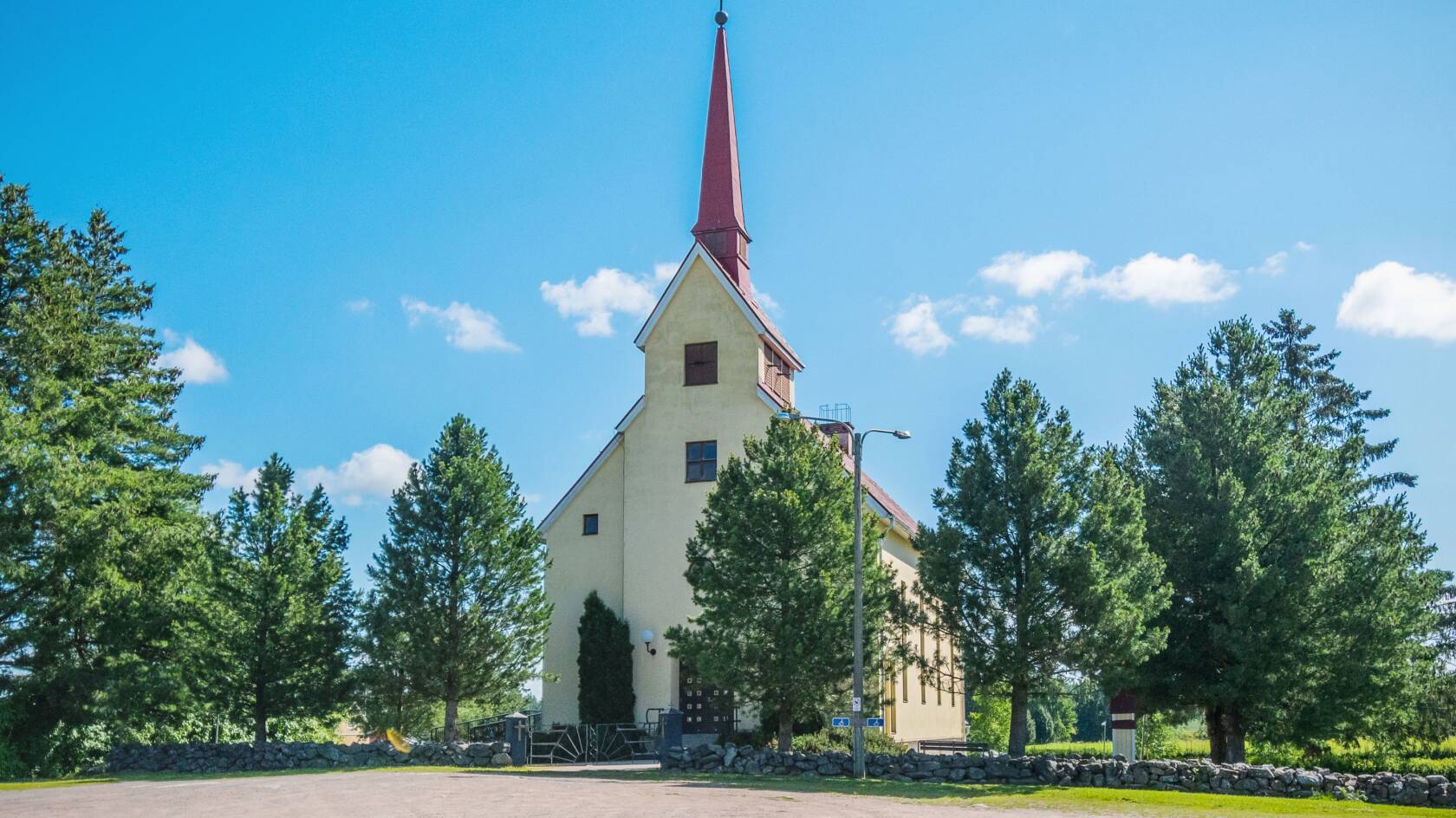 Vaalea kirkko, jonka ympärillä lehtipuita, nurmikko ja iso hiekka parkkialue.