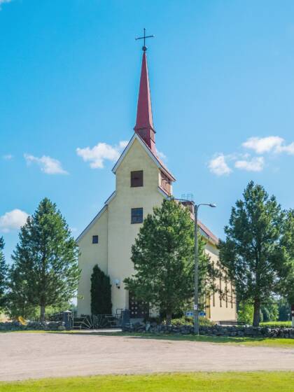 Vaalea kirkko, jonka ympärillä lehtipuita, nurmikko ja iso hiekka parkkialue.