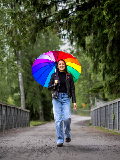 Nainen kävelee sillalla ja hänellä on kädessän sateenvarjo, jossa sateenkaaren värit.
