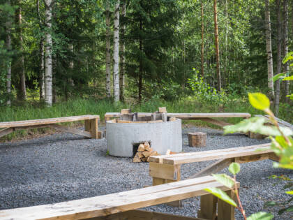 Pyöreä betoninen nuotiopaikka, ympärillä pitkät penkit, taustalla metsää.