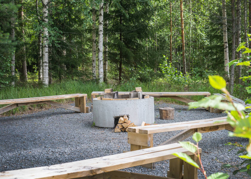 Pyöreä betoninen nuotiopaikka, ympärillä pitkät penkit, taustalla metsää.
