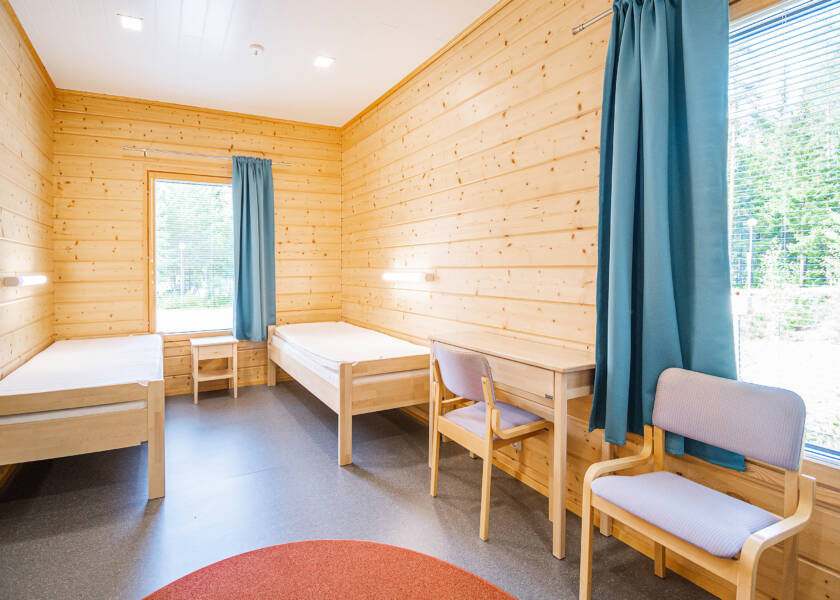 Hirsiseinäinen huone, jossa kaksi sänkyä seiniä vasten, pieni kirjoituspöytä ja kaksi tuolia.